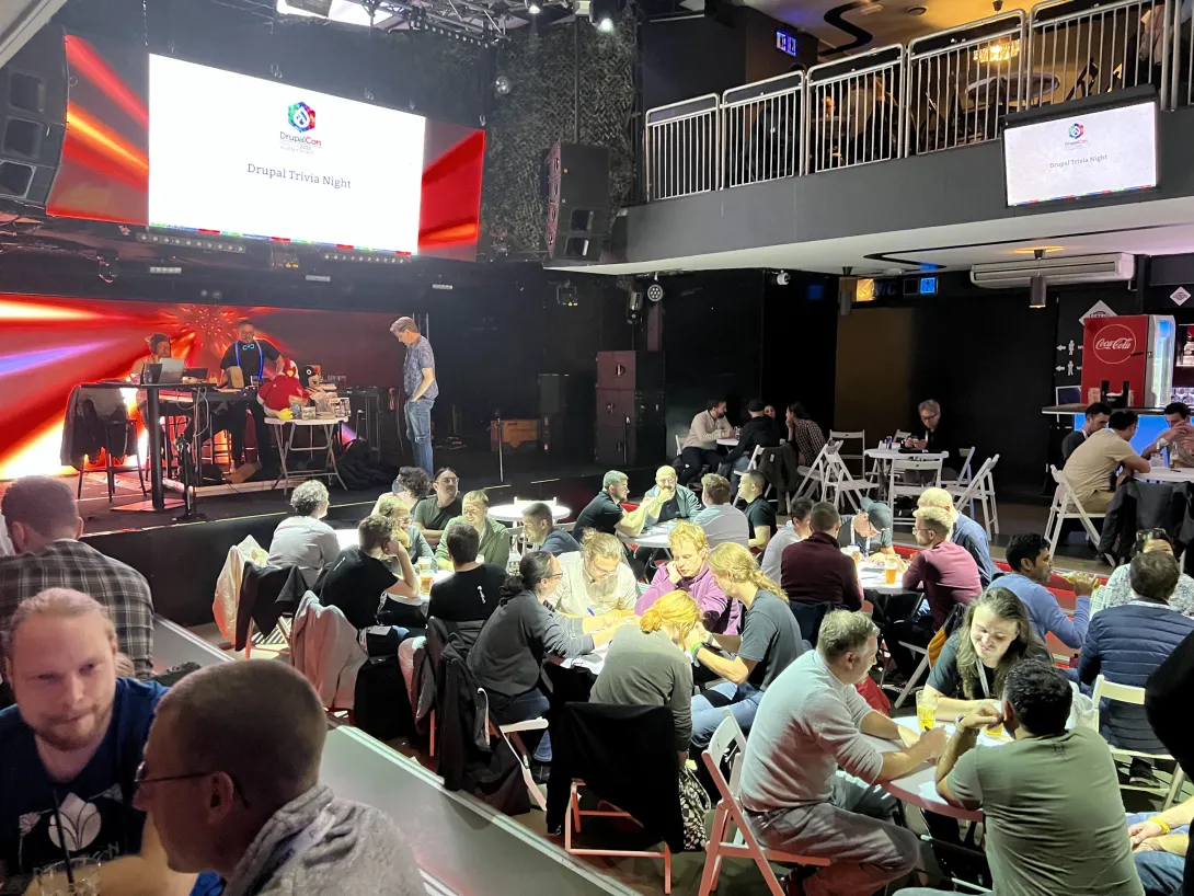 Groups of people around many tables at a music venue with low light and a banner that says "DrupalCon Trivia"