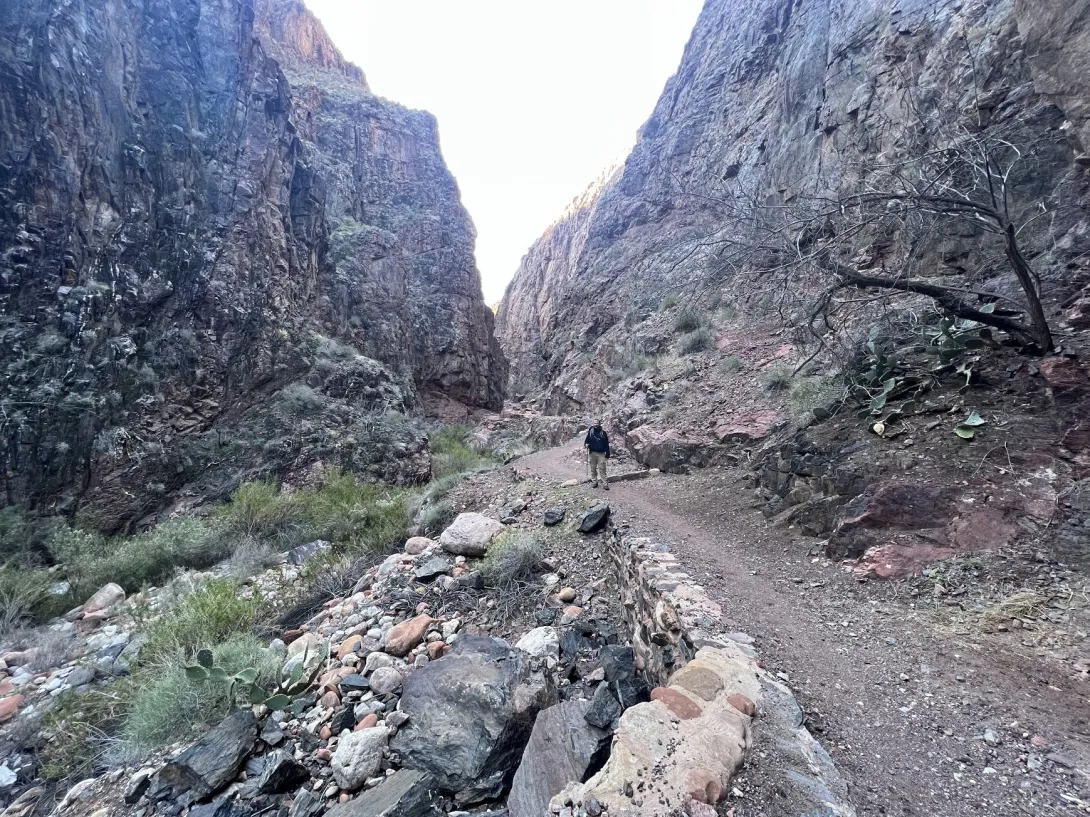 View of a trail down a narrow canyon with a hiker before. There's a small stream on the left