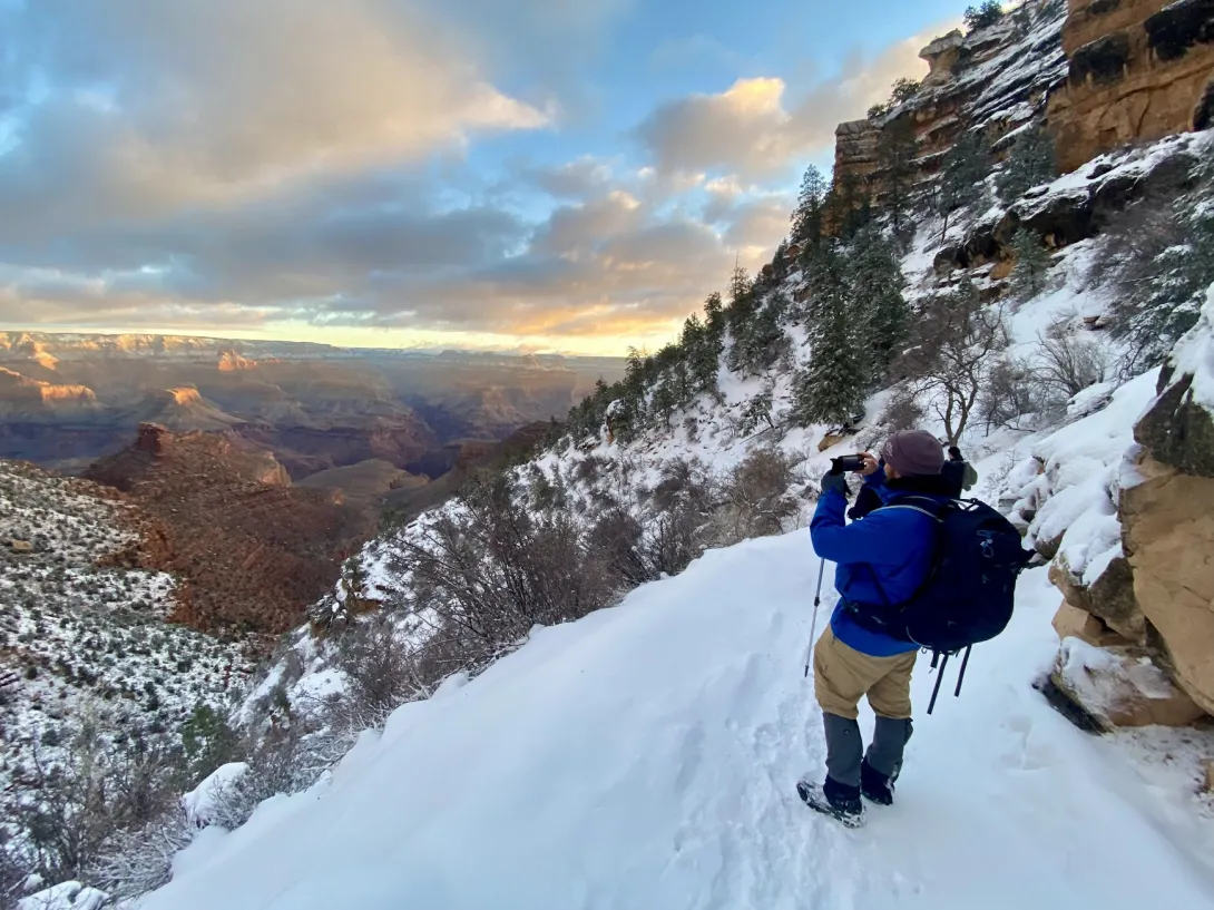 Me in a blue jacket and lots of gear taking a picture of the grand canyon on a steep snowy trail. The view of the canyon is majestic.