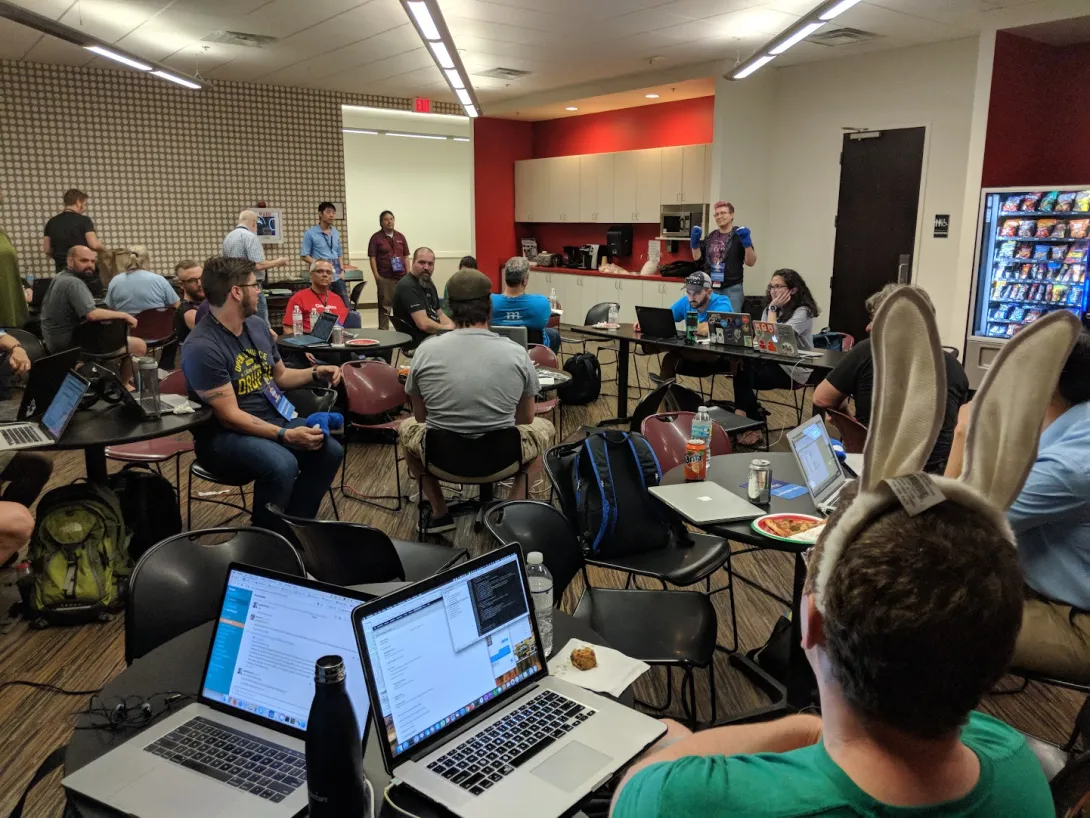 Lots of attendees sitting around tables with lots of computers. One attendee is speaking to everyone from the back