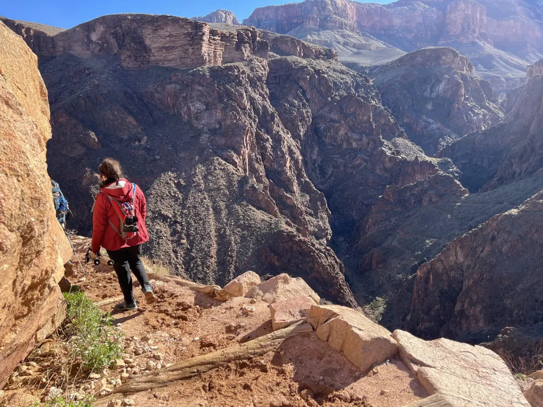 View of the trail overlooking a steep cliff. A hiker is going down the trail and you can see the canyon wall of the far side.