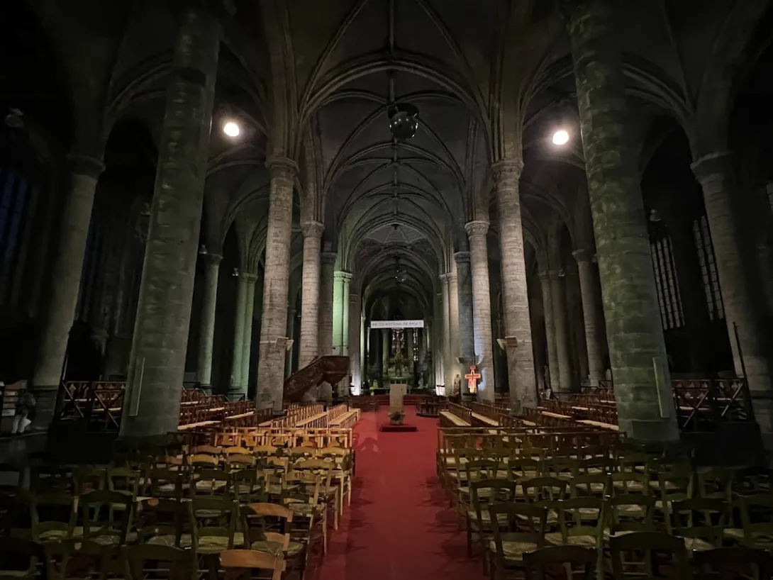 Inside of a large cathedral with lots of wooden chairs, red carpet, large columns. Very dimly lit.